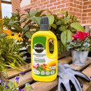 A yellow bottle of Miracle-Gro Performance Organics fertilizer sits on a wooden table surrounded by vibrant potted plants and gardening gloves, against a red-brick wall.