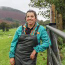 A person in teal and black farm attire leans on a metal fence, smiling. Behind them is a green field with sheep, and a misty hill in the background.