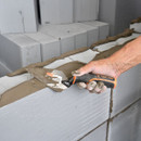 A hand uses a Tactix tool to spread mortar on a partially built wall of light grey concrete blocks. The scene conveys precision and craftsmanship.