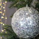 Close-up of a sparkling, silver sequin Christmas ornament hanging on a green tree branch with warm, blurred lights in the background. Festive and bright.