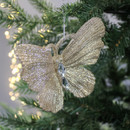 A glittery gold butterfly ornament with a sparkling, textured surface hangs on a green Christmas tree branch, surrounded by soft glowing lights.