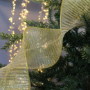Golden ribbon gracefully drapes over lush green pine branches, with soft twinkling lights in the background, evoking a festive holiday mood.