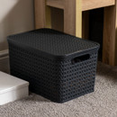 Grey woven plastic storage basket with lid placed on a carpeted floor beside a wooden side table, modern home décor storage solution for living room organisation.