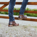 Close-up of a person in blue jeans and brown leather boots perched on a wooden fence outdoors. The scene conveys a rustic, relaxed vibe.