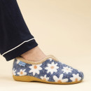 Side view of a foot wearing a cosy slipper with a blue fuzzy fabric covered in white and orange daisy patterns, trimmed with beige faux fur, and set against a beige background. The wearer is dressed in navy pyjamas with white piping.