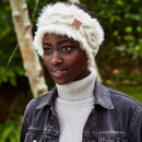 A person wearing a fluffy white headband and denim jacket smiles subtly amid lush greenery, exuding warmth and calm in an autumn setting.