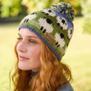 A woman with red hair smiles subtly, wearing a colourful knit hat with sheep patterns and a pompom. Sunlight filters through a green, blurred background.
