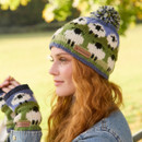 A person with long red hair wears a colourful wool hat and mittens featuring white sheep designs. The background is a sunlit park with green trees.