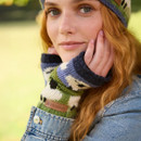 A person with red hair, wearing a denim jacket and colourful knit fingerless gloves, smiles softly. The background is a blurred green outdoor setting.