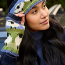 A woman with long dark hair wears a knitted blue, green, and white headband and matching fingerless gloves, gazing thoughtfully to the side.