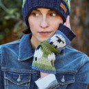 A woman in a denim jacket wears a knitted navy hat and multicoloured fingerless gloves, gazing thoughtfully. The background features blurred greenery.