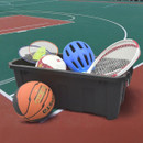 A black container on a sports court holds badminton rackets, a shuttlecock, a volleyball, blue helmet, and tennis rackets. A basketball rests beside it.