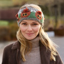 Smiling person outdoors wearing a knitted headband with red and green floral patterns. Dressed in a brown coat and grey turtleneck, with a blurred background. Smiling person outdoors wearing a knitted headband with red and green floral patterns. Dressed in a brown coat and grey turtleneck, with a blurred background.
