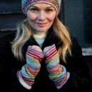 Woman smiling warmly, wearing a knitted hat and rainbow-striped fingerless gloves. She stands in front of a dark background, exuding a cosy, cheerful vibe.