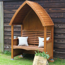 Stained beech wood cottage arbour bench with curved slatted roof and slatted back, styled with cushions beside a dark timber wall.