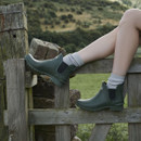 Green Barbour Chelsea wellington boots worn outdoors with grey socks, set against rolling hills and farmland.