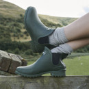Person wearing green Barbour Chelsea wellington boots while resting feet on a wooden fence in the countryside.