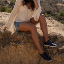 Woman in denim shorts and white blouse wearing navy lace-up leather shoes on sunlit rocks by the sea.