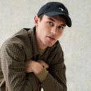 Young man in a checked shirt and black cap leans forward, arms crossed, gazing thoughtfully. Neutral background enhances his relaxed, contemplative expression.