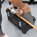A person uses a hand saw to cut a wooden plank on a black tool box, supported by their foot in a trainer. The scene conveys focus and precision.