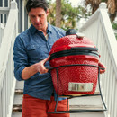 A person in a blue shirt and orange trousers carries a large red grill downstairs with greenery in the background.