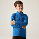 A smiling boy in a bright blue fleece crosses his arms, conveying a cheerful and relaxed mood. The background is a plain light gray.