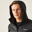 A person wearing a black hooded jacket gazes directly at the camera against a plain beige background, conveying a calm and confident demeanour.