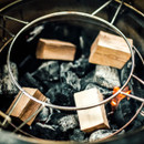 A close-up of glowing charcoal with wooden blocks arranged above, contained in a circular metal rack, ready for grilling.