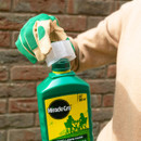 Gloved hand opening green bottle of Miracle-Gro liquid lawn food against brick wall background. The tone is practical, focused on gardening.