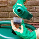 A person in green gardening gloves pours liquid into a measuring cup marked in milliliters, above a green watering can. The background is a brick wall.