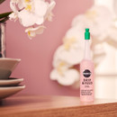 A pink bottle labelled "Drip & Feed" with a green cap sits on a wooden surface. White orchids in soft focus are in the background, creating a serene tone.