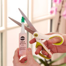 A hand holds a bottle of plant fertilizer next to scissors against a bright window backdrop with pink flowers, conveying a gardening theme.
