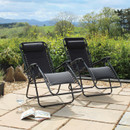 Two black reclining lounge chairs on a stone patio, surrounded by greenery and mountains under a partly cloudy sky. A magazine rests nearby.