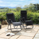Two reclining chairs on a stone patio, surrounded by greenery and mountains in the background, with a magazine and wine glass nearby.