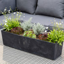 A rectangular black planter with green and purple succulents, small white flowers, and textured greenery on a woven surface, near grey cushions.