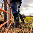 Person wearing tall black Muck Boot Muckmaster wellingtons standing by a farm gate in a muddy field.
