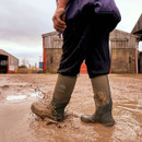 Person wearing green Muck Boot Arctic Sport boots walking through a muddy farmyard with water splashing around the boots. Person wearing green Muck Boot Arctic Sport boots walking through a muddy farmyard with water splashing around the boots.