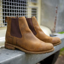 Brown leather Chelsea boots with dark elastic side panels rest on a metal surface outdoors. The rustic setting suggests durability and ruggedness.