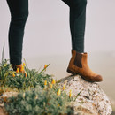 A person wearing brown boots and dark pants stands on a rocky surface surrounded by small yellow flowers, conveying a sense of adventure and nature.