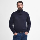 A man with a beard in a navy zip-up jumper and jeans stands confidently with hands in pockets against a plain white background. Relaxed and casual vibe.