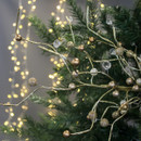 Close-up of a festive Christmas decoration with gold branches and ornaments, set against a backdrop of glowing fairy lights and green pine needles.
