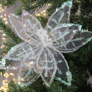A delicate white poinsettia ornament glistens with sequins on a green Christmas tree, surrounded by soft, blurred twinkling lights. Holiday ambiance.