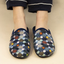Person wearing slip-on Lunar slippers with a cosy knit-like upper in multi-coloured patchwork and scattered pastel polka dots, standing on a neutral background while dressed in navy pyjama trousers with white piping.