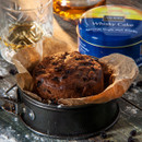 A rich, dark whisky cake in a baking tin with parchment paper sits on a rustic table. A whiskey glass and a blue tin labelled "Whisky Cake" are nearby.