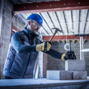 Construction worker wearing blue safety helmet, protective goggles, and gloves using hammer and chisel on concrete block inside industrial building site with exposed beams