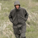Man wearing a hooded dark green waterproof jacket and outdoor trousers while walking through grassy countryside.