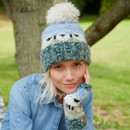 A person in a denim jacket smiles softly, wearing a knit hat and gloves with sheep patterns. Background shows a tree and green grass, creating a cosy, rustic feel.