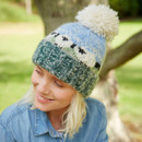 A person smiles outdoors, wearing a blue and green knitted hat with a large pom-pom. The background shows a tree and grassy field, conveying a relaxed, cheerful mood.