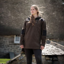 A woman stands confidently outdoors in front of a rustic stone building. She wears a brown jacket, with wind gently blowing her hair. The scene feels serene and resilient.