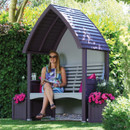 Lavender and stone painted garden arbour under trees, with a woman seated on the bench and matching side planters filled with bright pink flowers.